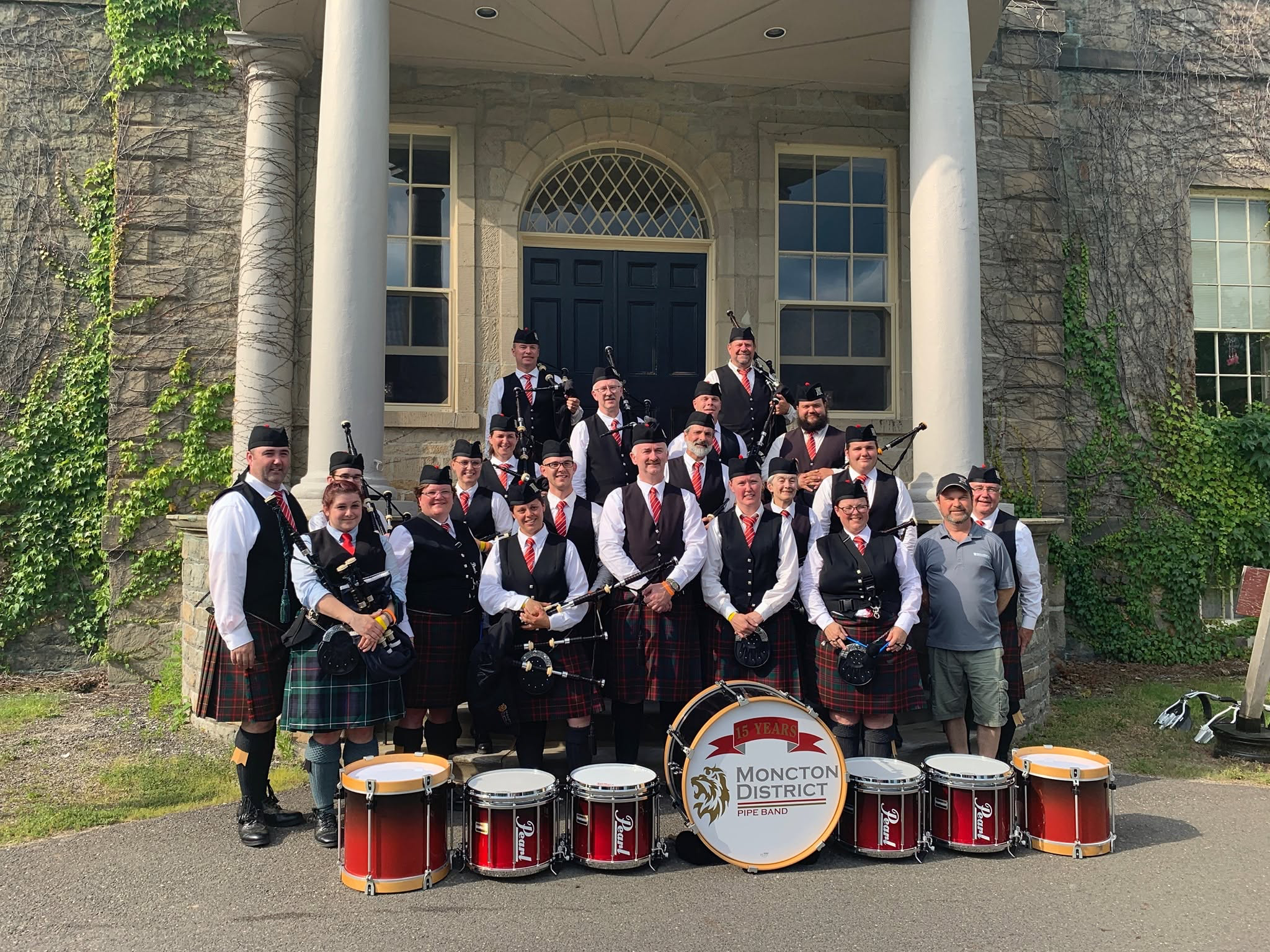 Moncton District Pipe Band gathered with pipes and drums outside a stone building